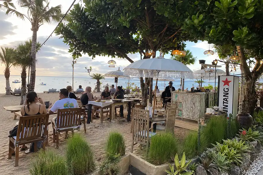 People dining at open-air beachfront tables under white ceremonial umbrellas at dusk.