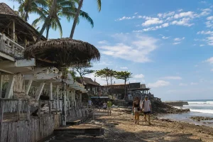 People walking along a sandy shoreline lined with rustic wooden beach cafes under a blue sky.