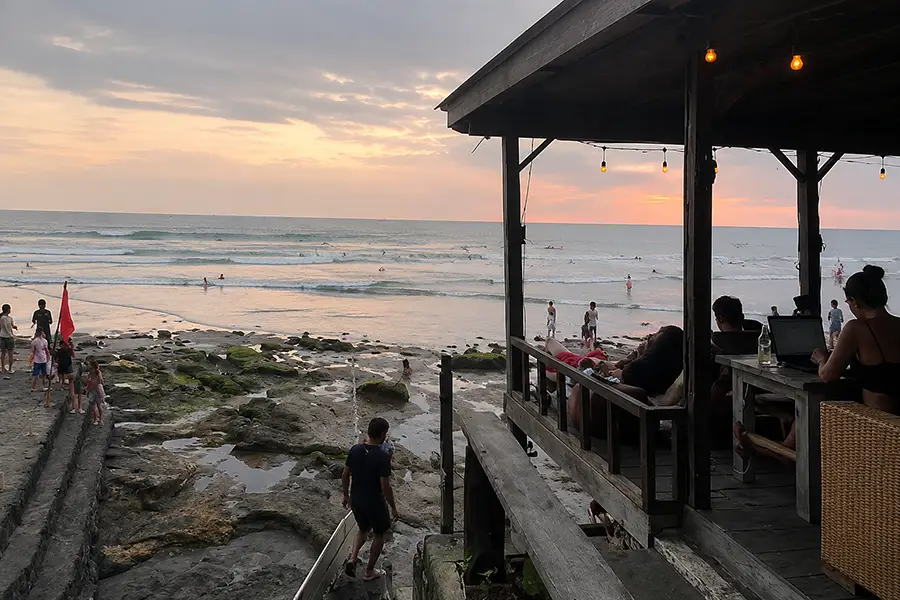 People relaxing in a wooden beachside hut overlooking surfers and rocky tide pools during sunset.