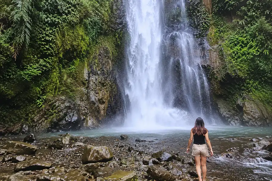 A traveler standing near rocky shallow water looking up at a massive cascading waterfall.