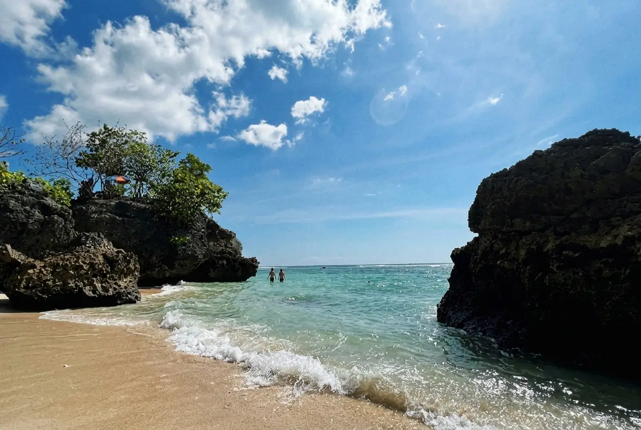 People swimming in clear turquoise water framed by massive rock formations under a bright blue sky.