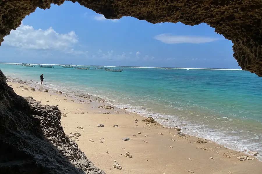 View from inside a dark limestone cave looking out at a sunny turquoise beach with boats floating in the water.