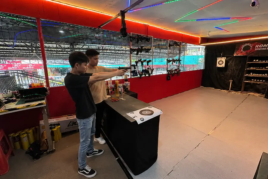 Two men standing in an indoor shooting bay practicing handgun aiming next to wall-mounted airsoft guns.