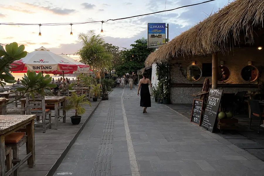 A woman walking along a paved coastal path lined with beachside cafes and thatched-roof restaurants at dusk.