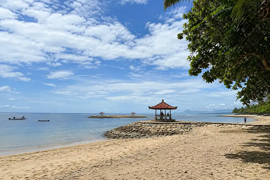 A traditional wooden gazebo sitting at the end of a stone breakwater on a quiet beach.