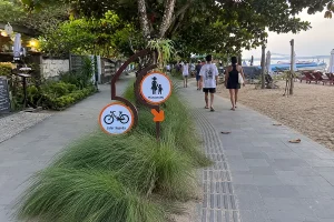Tourists strolling past orange pedestrian and bicycle lane signs on a paved beachfront path.