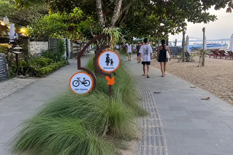 Tourists strolling past orange pedestrian and bicycle lane signs on a paved beachfront path.