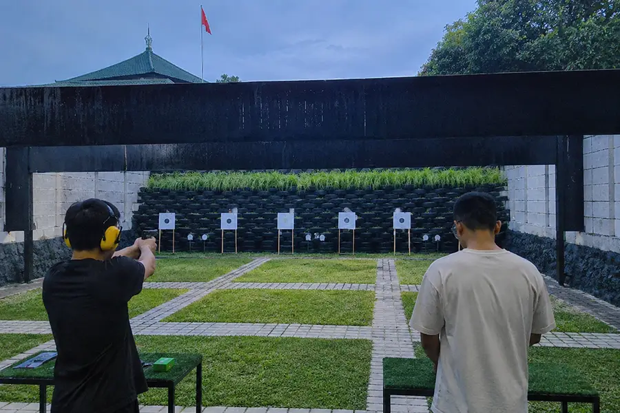 Two men practicing target shooting at an outdoor range with green grass and paper targets.