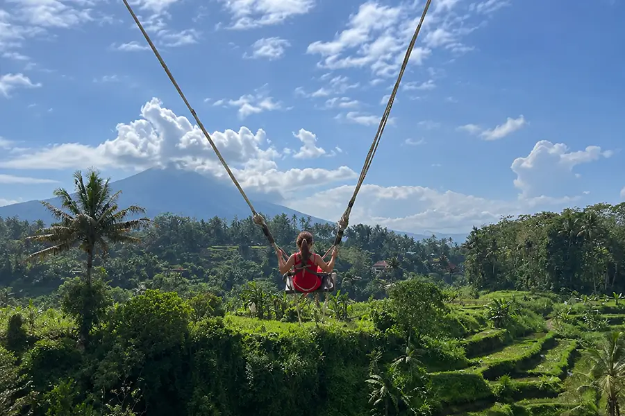 A woman riding a high swing over lush fields facing Mount Agung at Maha Gangga Valley.