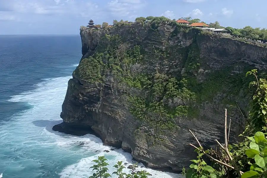 A traditional Balinese temple perched on top of a steep ocean cliff overlooking crashing blue waves.
