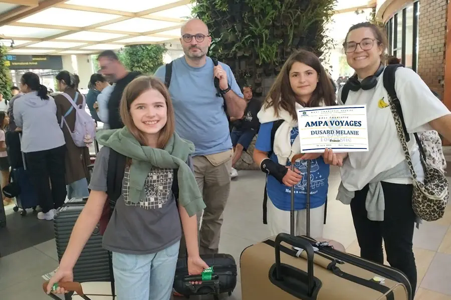 A smiling tourist family holding a welcome sign with their luggage inside the airport terminal.