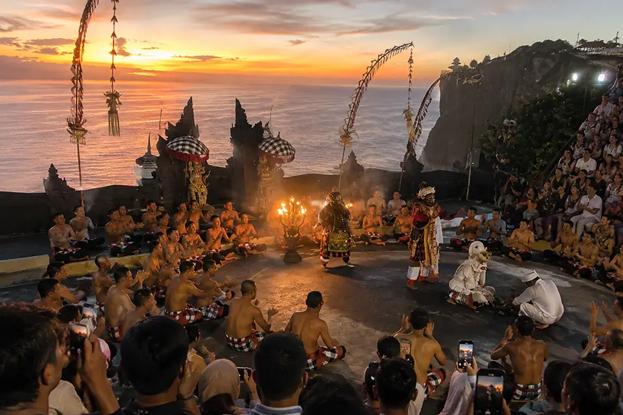 Performers conducting a Kecak fire dance in an open-air amphitheater on a cliff during sunset.